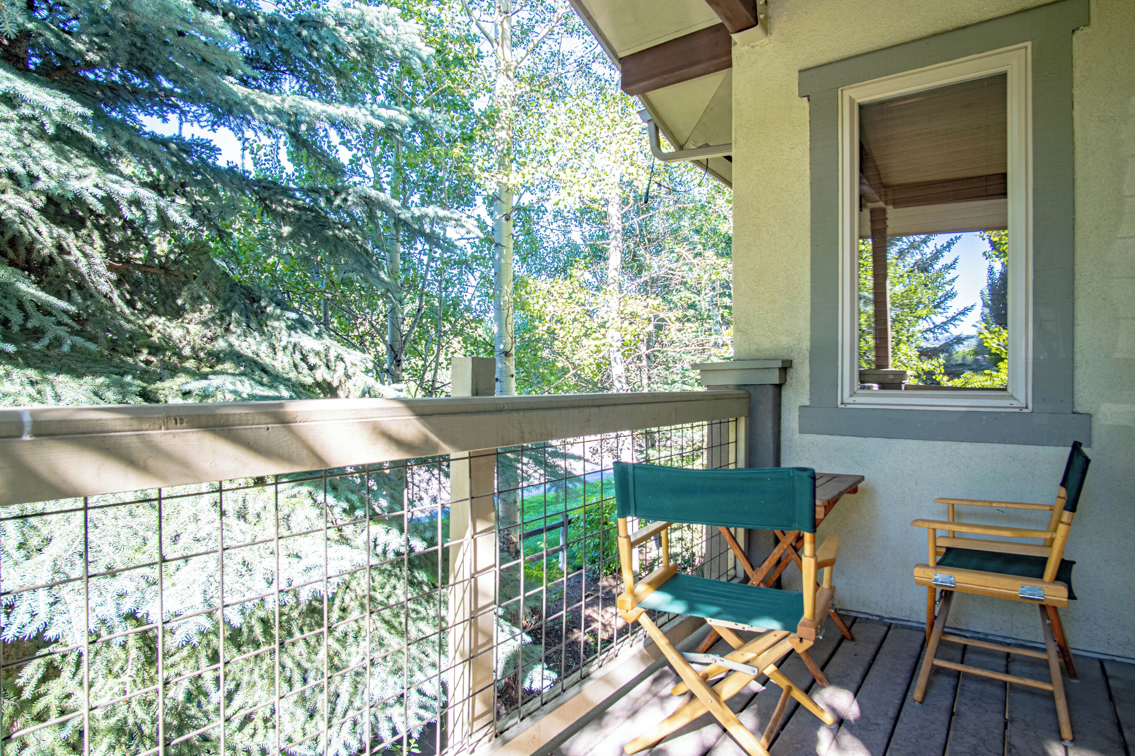 A balcony with a wooden railing, two green folding chairs, and a window overlooking a forest.