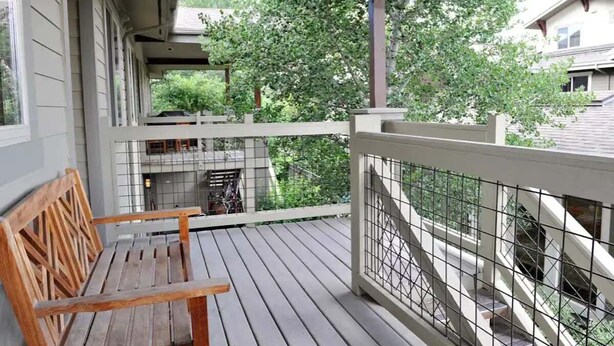 Um banco de madeira em um deck de madeira com guarda-corpo metálico, com vista para uma área residencial com árvores e casas.