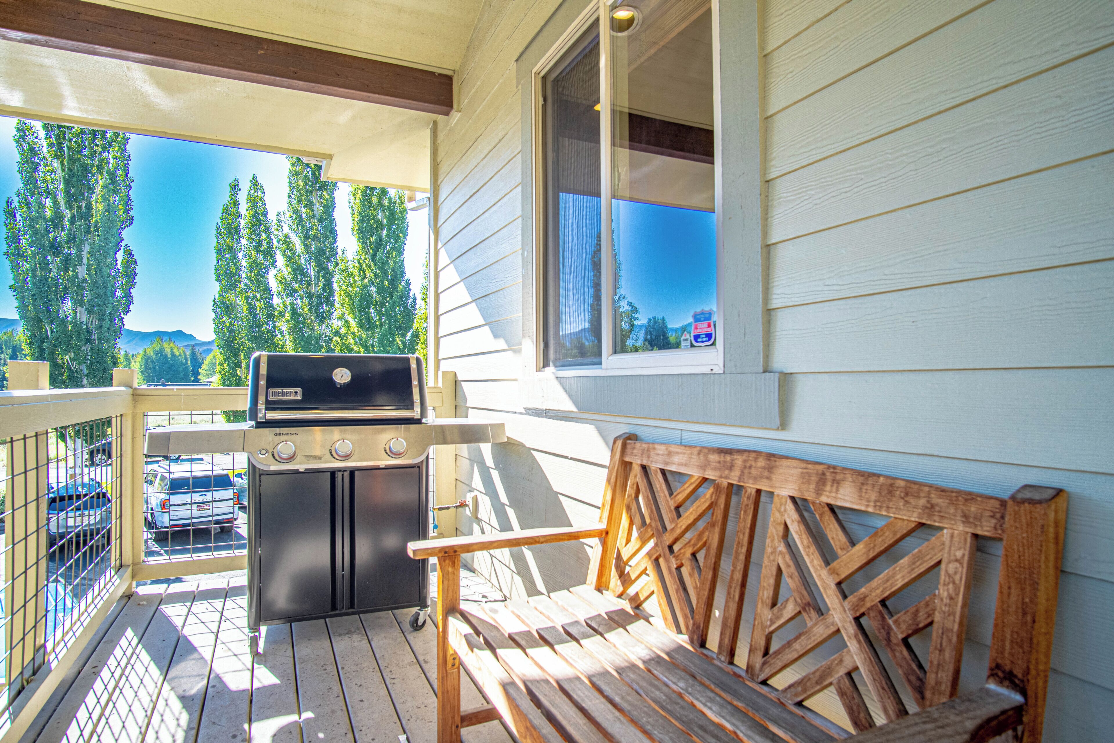 A wooden bench and a grill on a porch with a view of trees and a mountain.