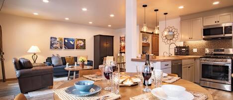 A modern living room with a round wooden dining table set for a meal, a sofa, and a kitchen area with stainless steel appliances.