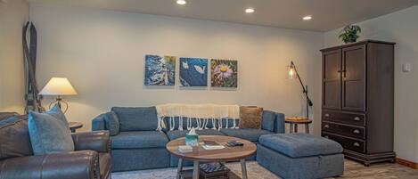 A living room with a sofa, armchair, ottoman, coffee table, lamp, cabinet, and artwork on the wall.
