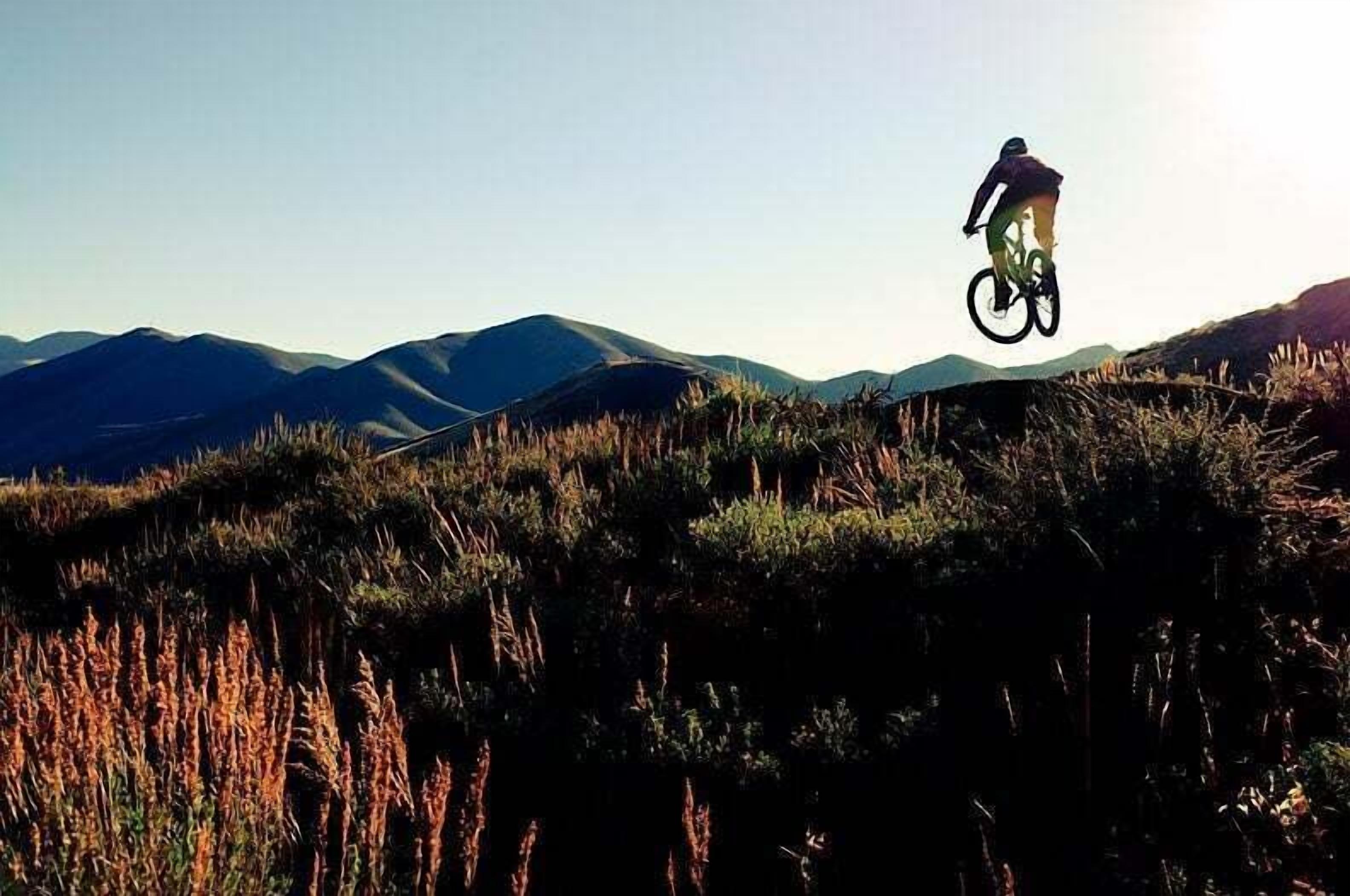A person on a mountain bike performing a jump over a grassy hill with mountains in the background.