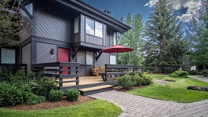 Une maison moderne à deux étages, avec une façade sombre, une porte rouge et une terrasse en bois surmontée d’un parasol rouge.