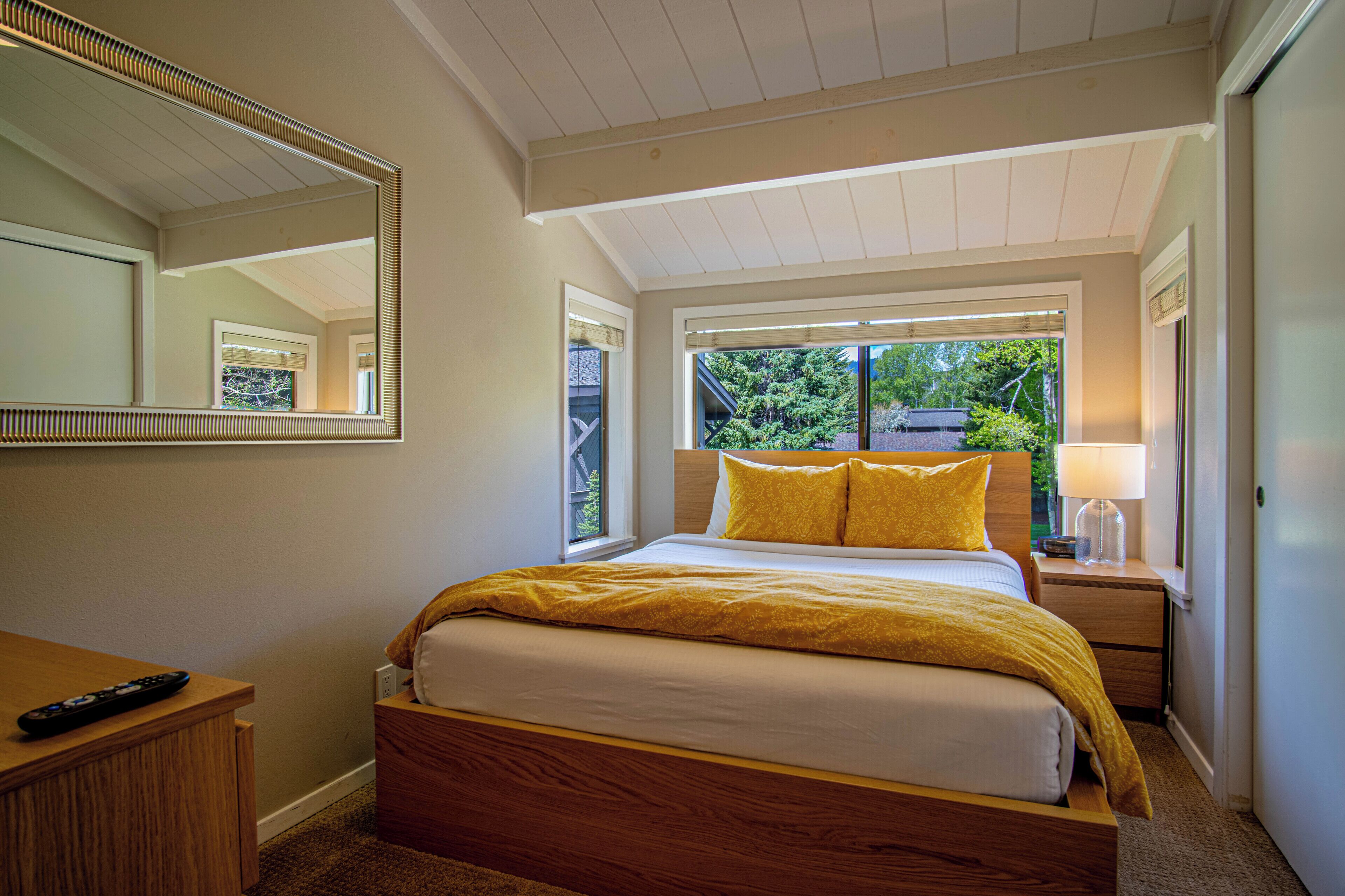 A bedroom with a bed, a nightstand, a lamp, a mirror, and a window with a view of greenery outside.