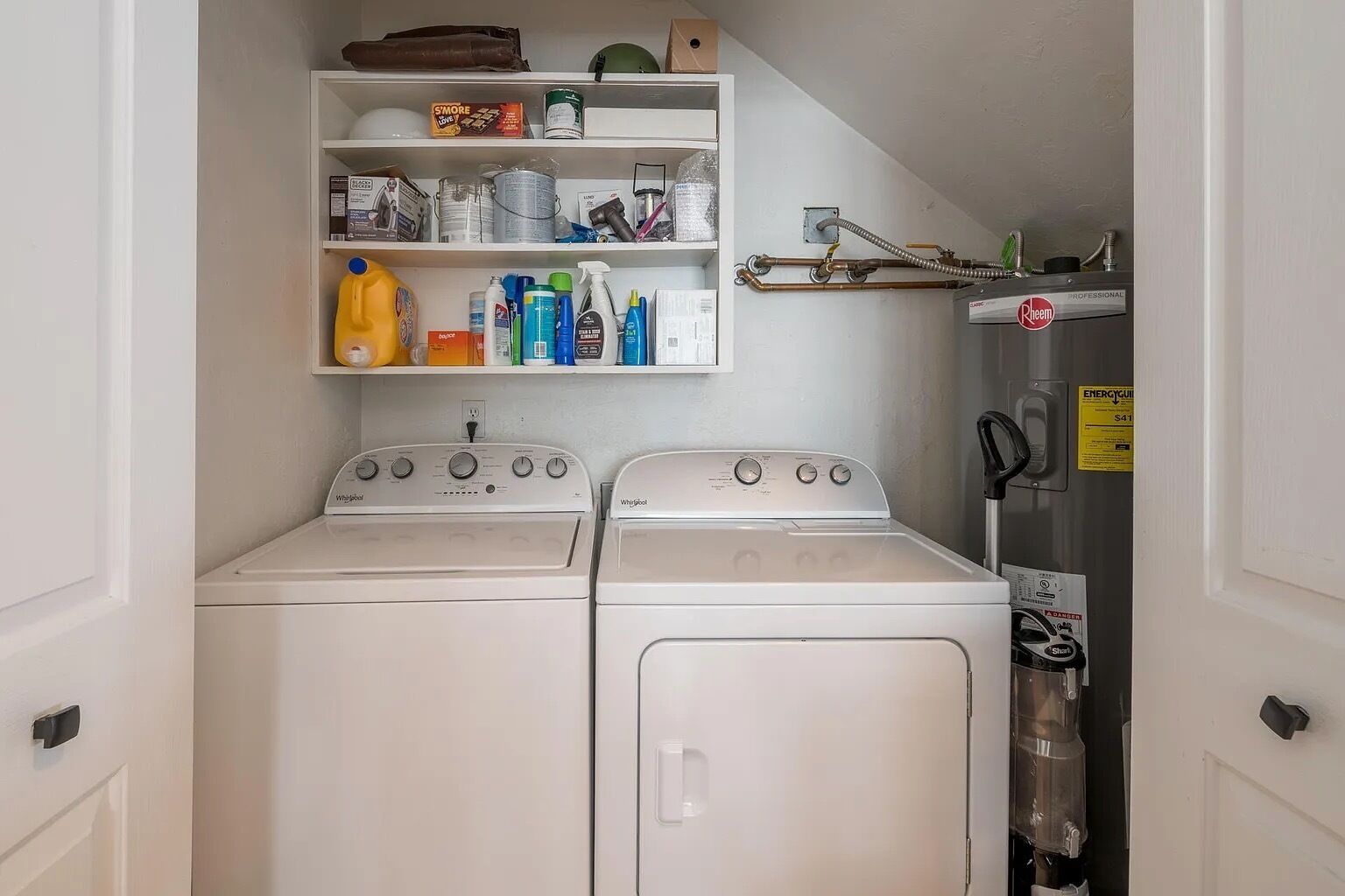 A laundry room with two Whirlpool washers and dryers, a shelf above containing cleaning supplies, and a water heater with an energy efficiency label.
