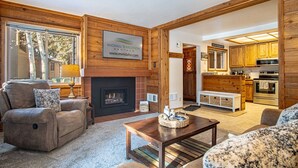 A cozy living room with a fireplace, a sofa, a coffee table, and a kitchen visible through an open doorway. - Elkhorn Bluff Condo + Complex & Resort Amenities (Sun Valley)