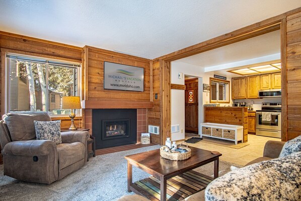 A cozy living room with a fireplace, a sofa, a coffee table, and a kitchen visible through an open doorway. - Elkhorn Bluff Condo + Complex & Resort Amenities (Sun Valley)