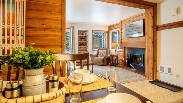 A cozy dining area with a wooden table set for two, a fireplace, and a view of a living room with a TV. - Elkhorn Bluff Condo + Complex & Resort Amenities (Sun Valley)