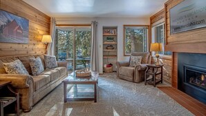 A cozy living room with a leather sofa, a recliner, and a fireplace. There are wooden shelves with decorative items and a large window offering a view of the outdoors. - Elkhorn Bluff Condo + Complex & Resort Amenities (Sun Valley)