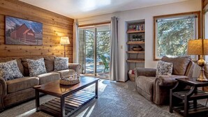 A cozy living room with a wooden wall, a large window, and a sliding glass door leading to an outdoor area. - Elkhorn Bluff Condo + Complex & Resort Amenities (Sun Valley)