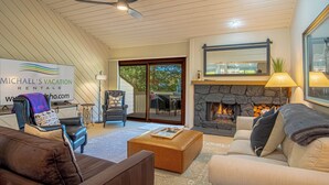 A spacious living room with a stone fireplace, a ceiling fan, and a sign for Michael's Vacation Rentals. - Ridge Condo - Luxury Condo W/resort Amenities (Sun Valley)