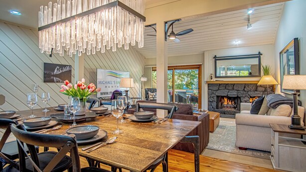 A dining area with a wooden table set for six, surrounded by chairs. A chandelier hangs above, and a stone fireplace is visible in the background. - Ridge Condo - Luxury Condo W/resort Amenities (Sun Valley)