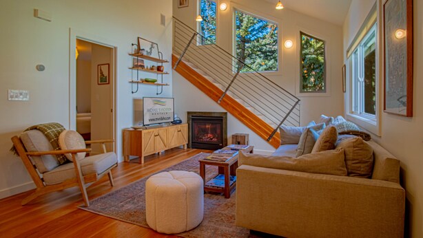 A spacious living room with a wooden staircase, a fireplace, and large windows offering views of trees. - 2bd, 2BA Ketchum Condo Newly Remodeled & Central (Ketchum)