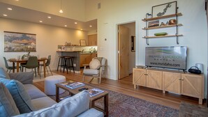 A modern living room with a sofa, coffee table, and a TV on a wooden cabinet. There is a dining area with chairs and a table, and a kitchen in the background. - 2bd, 2BA Ketchum Condo Newly Remodeled & Central (Ketchum)