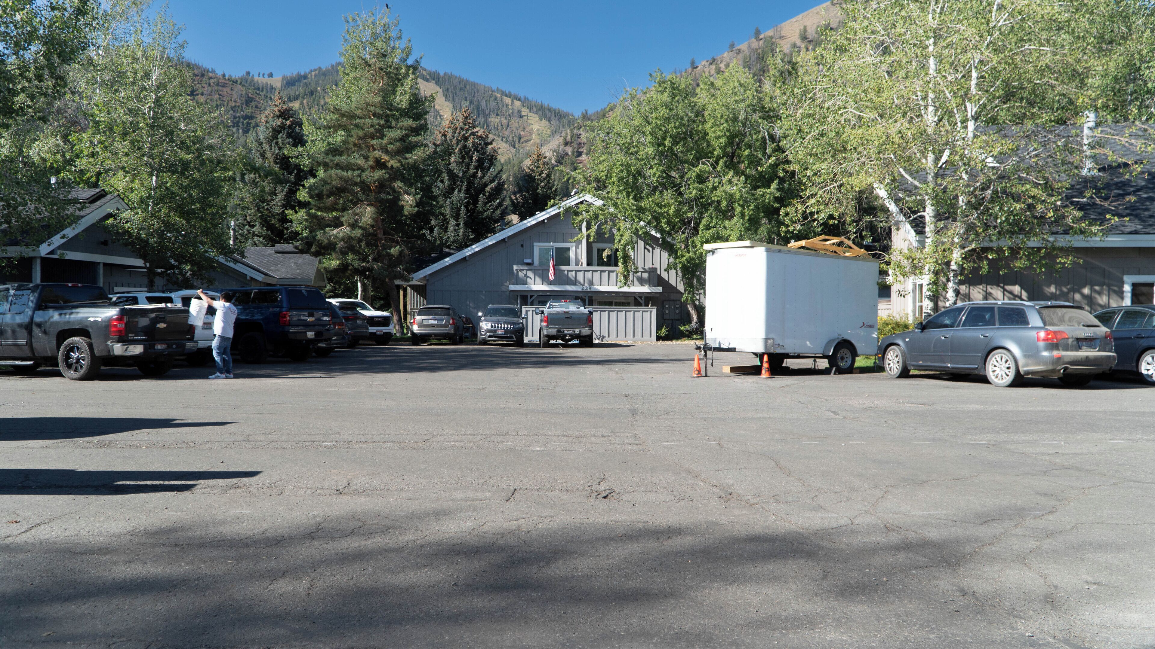 Un parking où sont stationnés plusieurs véhicules, dont des camions et une remorque blanche, devant un bâtiment doté d’un toit en pente et sur lequel flotte un drapeau.