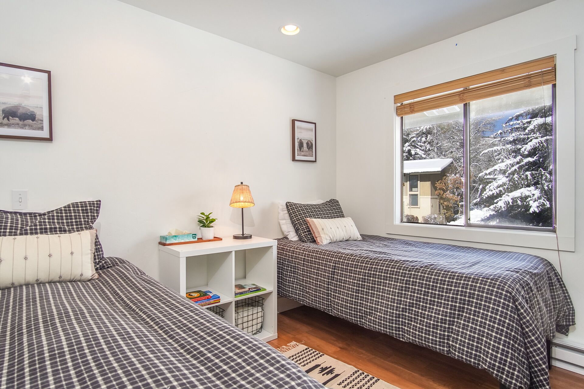 A bedroom with two beds, a bedside table, a lamp, a small shelf with books, and a window with a snowy view.