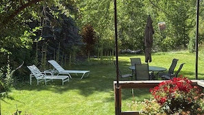A patio with a table and chairs, a red umbrella, and a garden with a lawn and trees. - Bigwood Bliss - 3BD Riverfront Home (Ketchum)