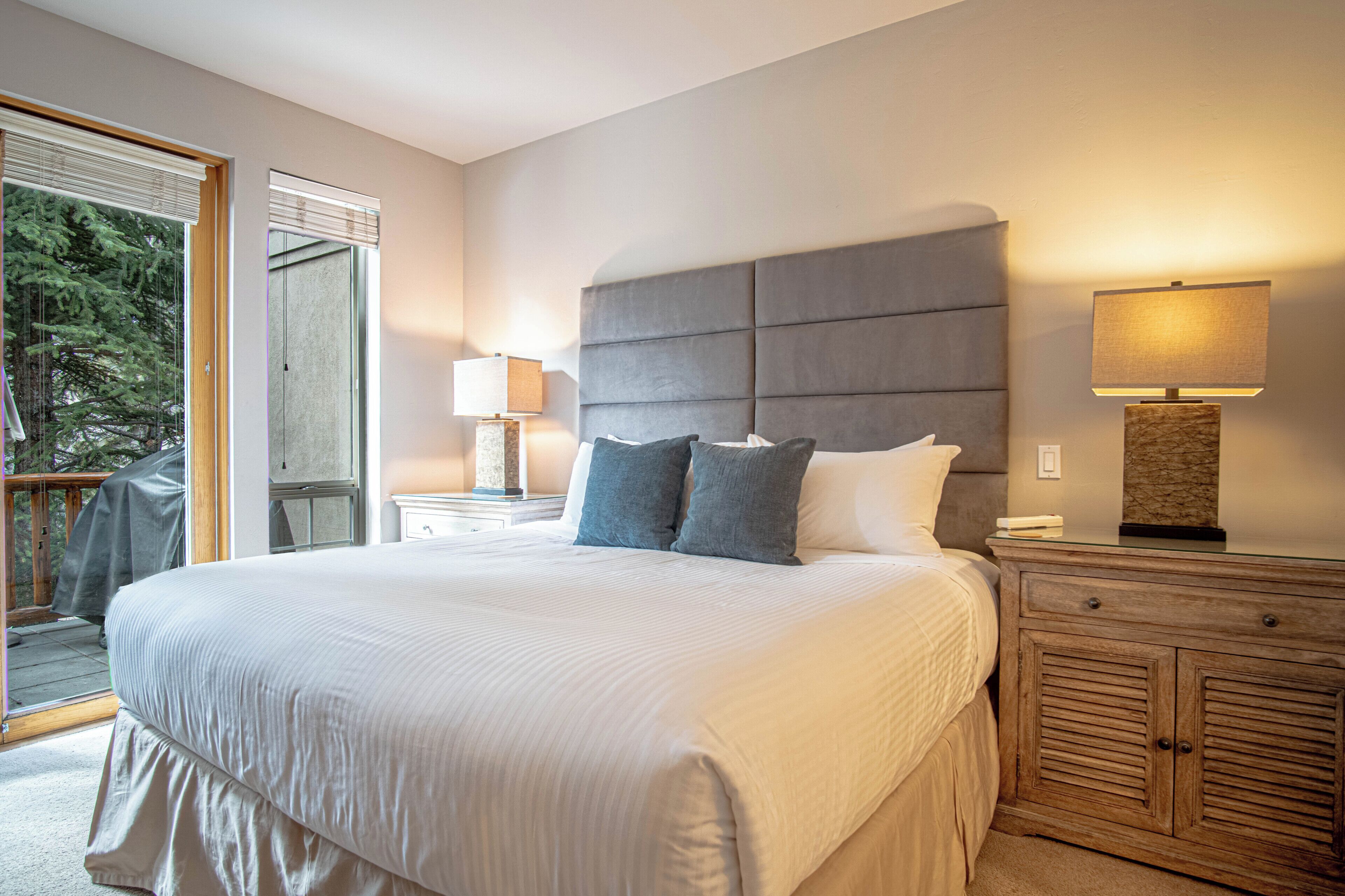 A bedroom with a large bed, two bedside tables with lamps, a wooden nightstand, and a sliding glass door leading to an outdoor area.