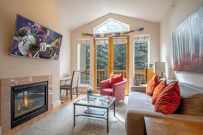 A living room with a fireplace, a sofa, a coffee table, and a painting of a football game. - Snowstar Hideaway - Hot Tub, Gym, Walk to Ski Lift (Ketchum)