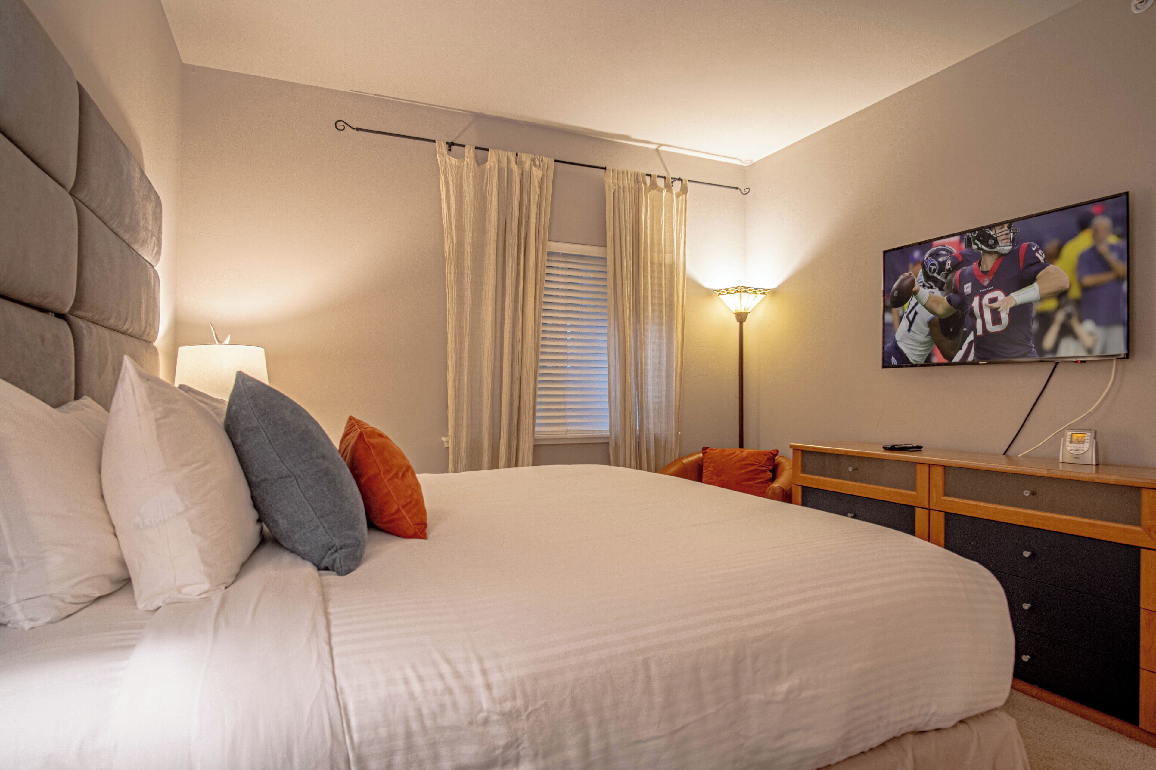 Un dormitorio con cama, almohadas, televisor, lámpara y ventana con cortinas.