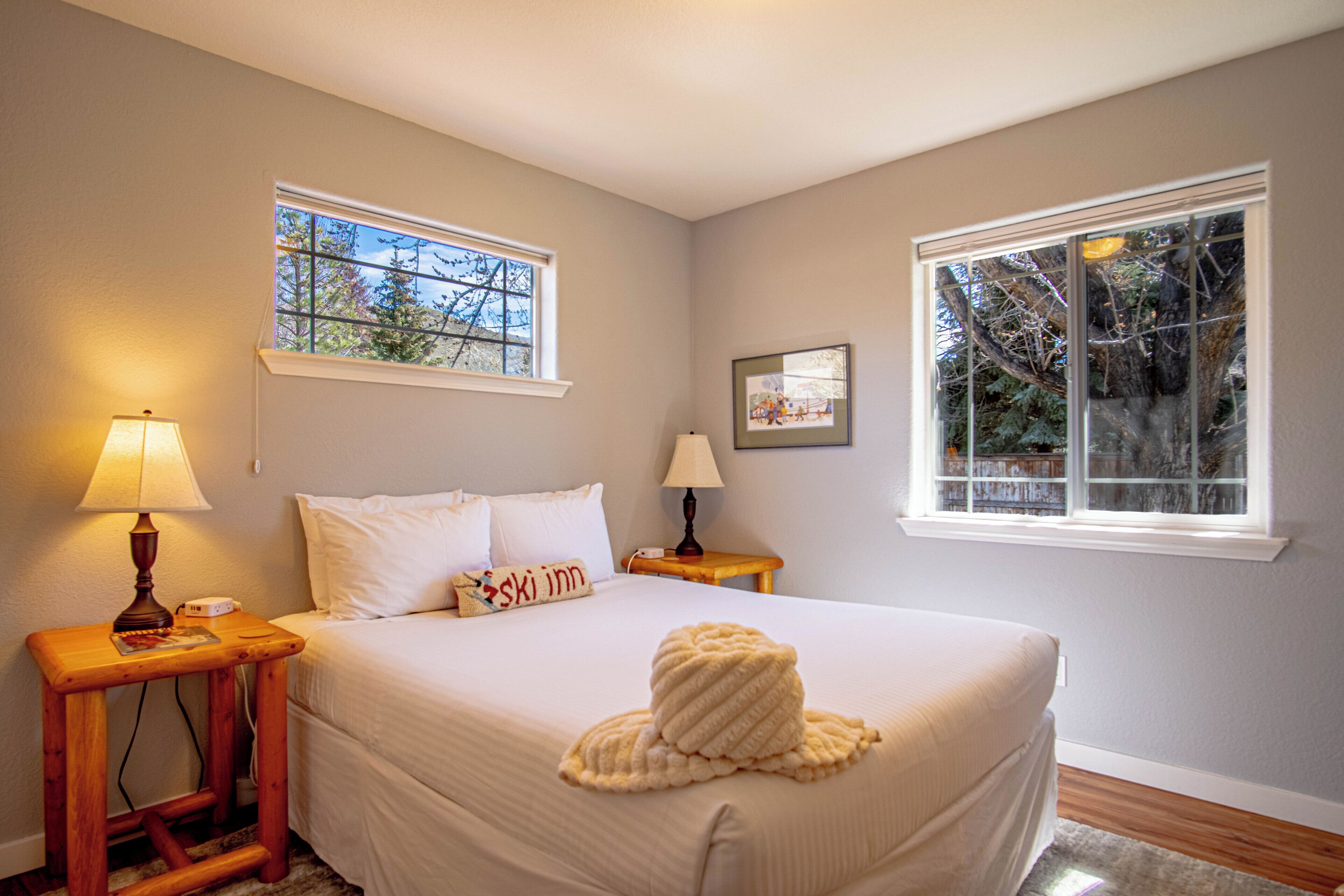 Un dormitorio con una cama, dos mesitas de noche con lámparas, una ventana con vista a árboles y un cuadro en la pared.