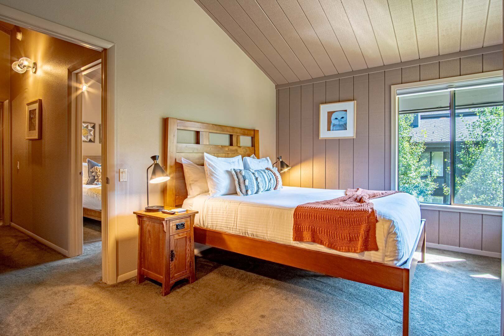 A bedroom with a wooden bed, a nightstand, a window with a view of greenery, and a door leading to another room.