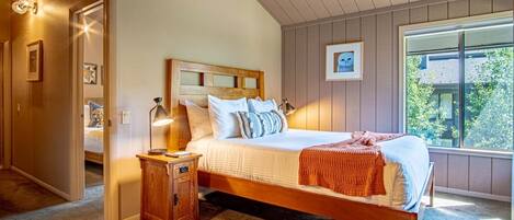 A bedroom with a wooden bed, a nightstand, a window with a view of greenery, and a door leading to another room.