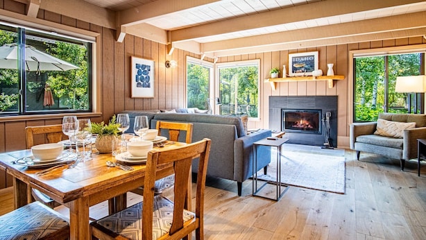 A cozy living room with a fireplace, wooden floors, and large windows offering a view of greenery. - Wildwood Cottage at Villager + SV Resort Amenities (Sun Valley)