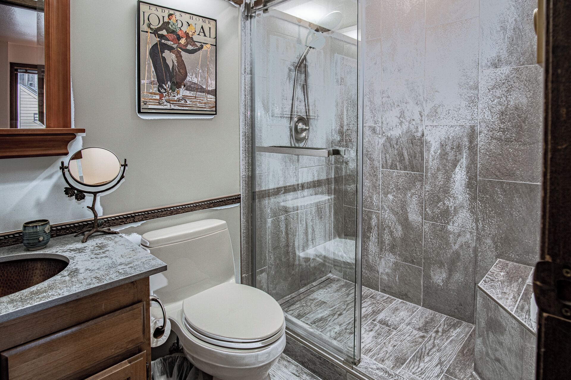 A bathroom with a glass shower enclosure, a toilet, a vanity with a mirror, and a wall-mounted picture.