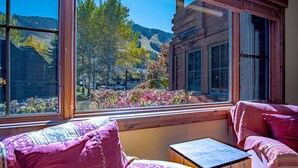 A room with large windows offering a view of trees and a mountain. There is a wooden table and two chairs with patterned cushions. - Lux Baldy Vista at Warm Springs Home + Hot Tub - Walk to SV Ski Lifts (Ketchum)