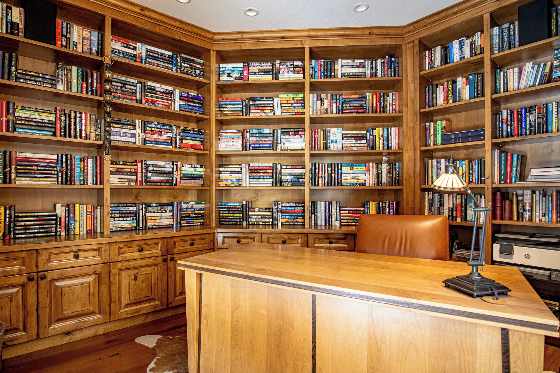 A library with wooden bookshelves filled with books, a desk with a lamp, and leather chairs.
