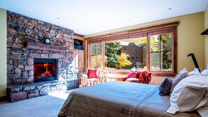 A bedroom with a stone fireplace, a large bed with multiple pillows, and a view of trees through large windows. - Lux Baldy Vista at Warm Springs Home + Hot Tub - Walk to SV Ski Lifts (Ketchum)