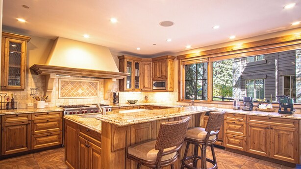 A spacious kitchen with wooden cabinets, a large island with bar stools, and a view of the outdoors through large windows. - Lux Baldy Vista at Warm Springs Home + Hot Tub - Walk to SV Ski Lifts (Ketchum)