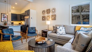 Un salon moderne avec un canapé gris, des fauteuils bleus et un pouf jaune. On aperçoit une cuisine à l’arrière-plan, avec des meubles de couleur sombre et un réfrigérateur en acier inoxydable.