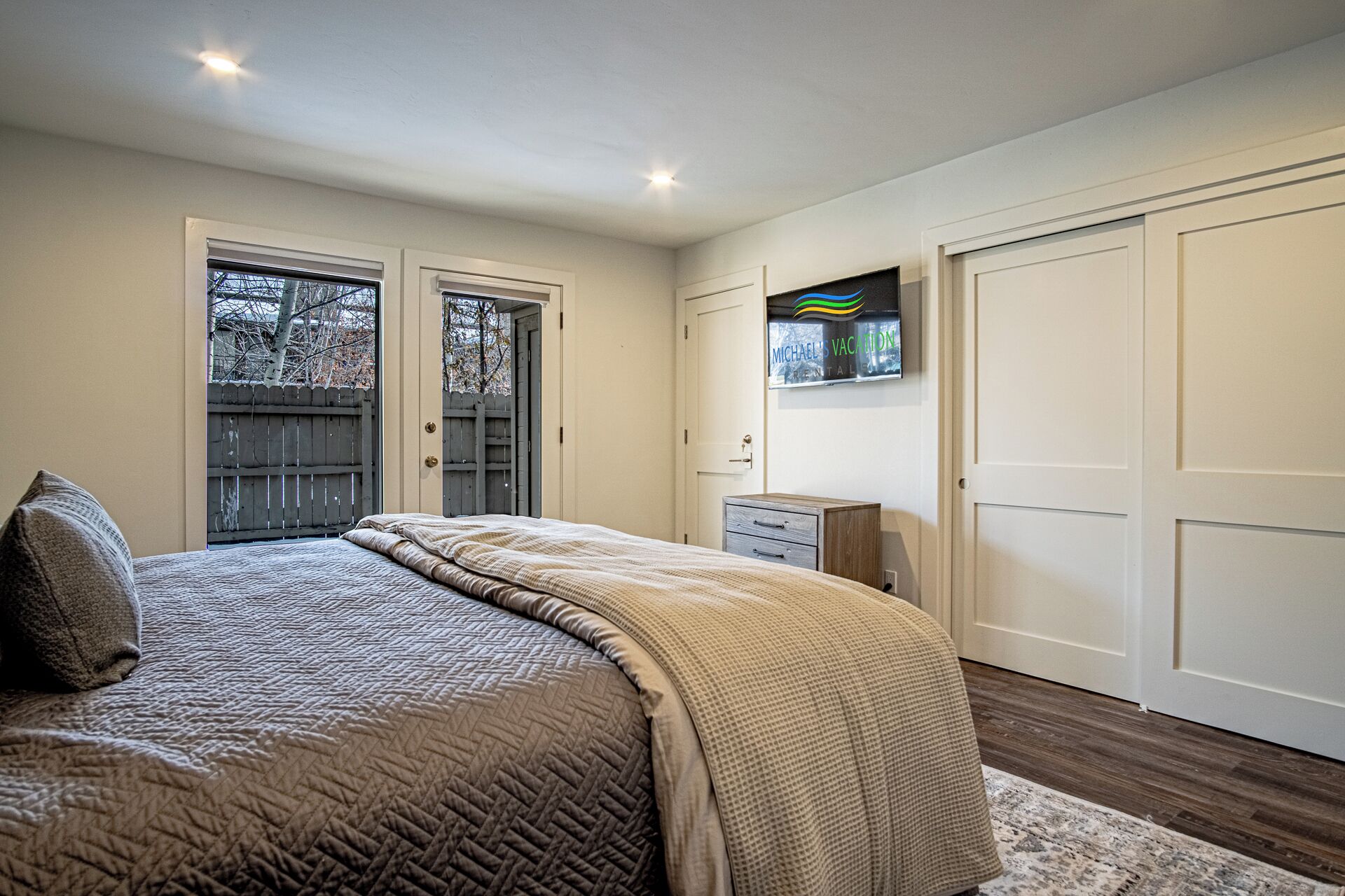 Une chambre à coucher avec un lit, deux portes, une petite armoire et une fenêtre donnant sur des arbres.