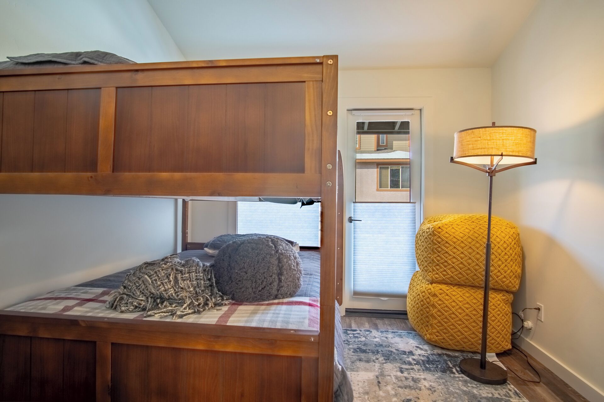 A bunk bed with a checkered blanket and a woven rug, a yellow woven ottoman, and a floor lamp in a room with a door leading to a balcony.