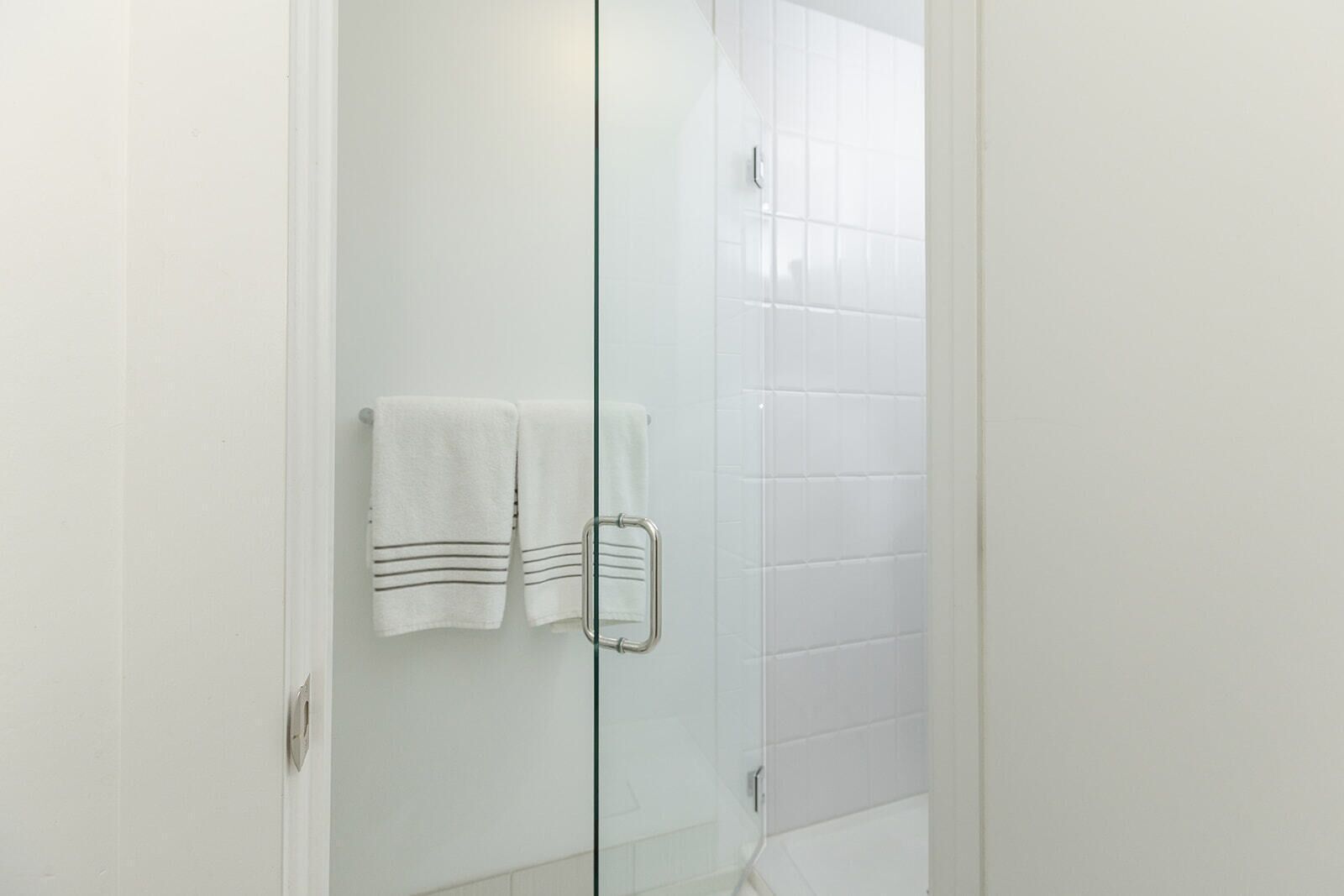 Una puerta de vidrio da acceso a un baño con azulejos blancos y dos toallas colgadas en un perchero.