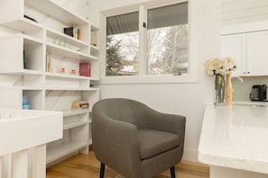 A modern kitchen with white cabinetry, a grey upholstered armchair, and a window with a view of bare trees. - Sunburst in Sun Valley! Modern Family Getaway in Elkhorn (Sun Valley)