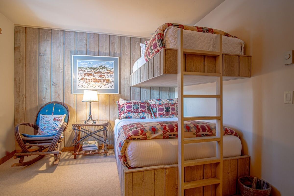 A bunk bed with a wooden headboard, a rocking chair, a small table with books, and a framed picture on the wall.