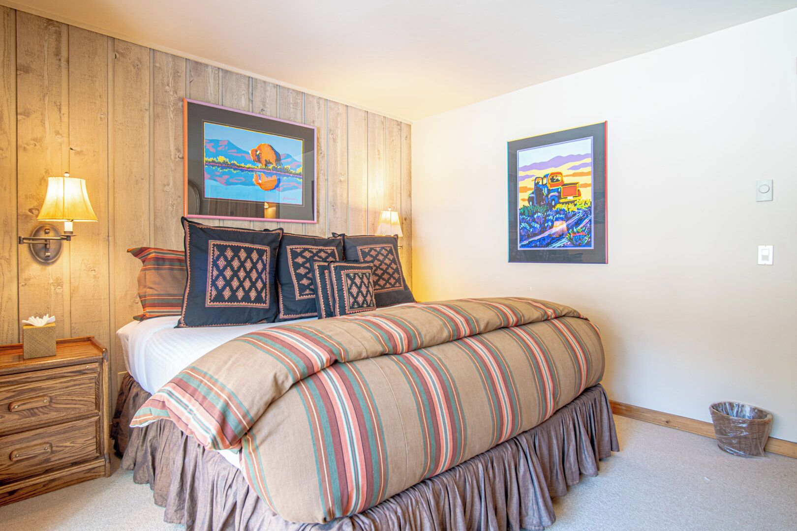 A bedroom with a wooden headboard, a bed with striped bedding, two wall-mounted lamps, and two framed paintings.