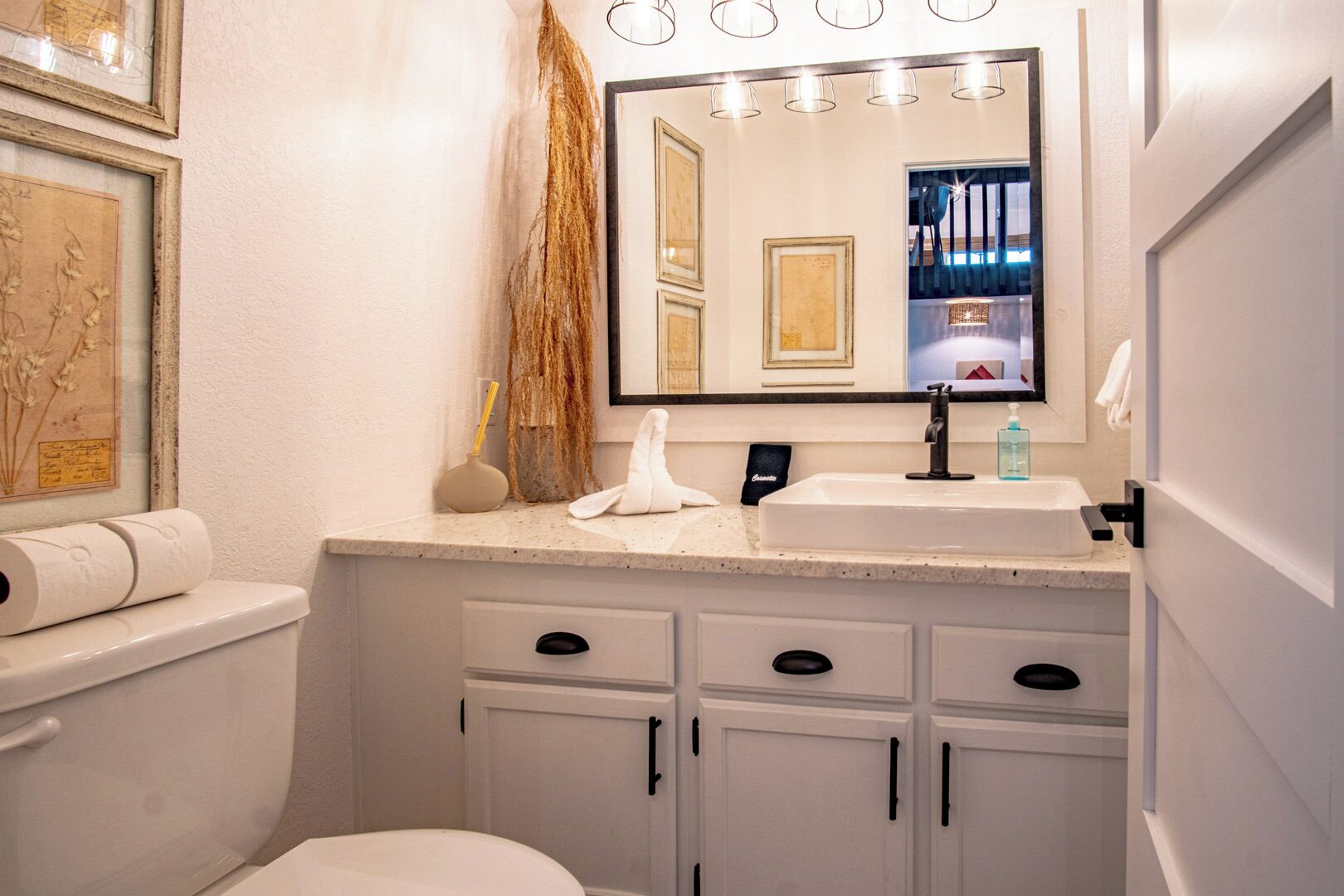 Une salle de bain avec un lavabo blanc, un miroir et des toilettes. Des tableaux encadrés ornent le mur et un rouleau de papier toilette est posé sur le comptoir.