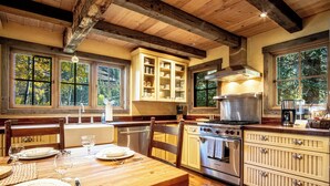 A wooden dining table set with plates, glasses, and cutlery, flanked by wooden chairs. A kitchen with wooden cabinets and a stove is visible in the background. - Board's Sunset Ranch - Sawtooth National Forest (Ketchum)