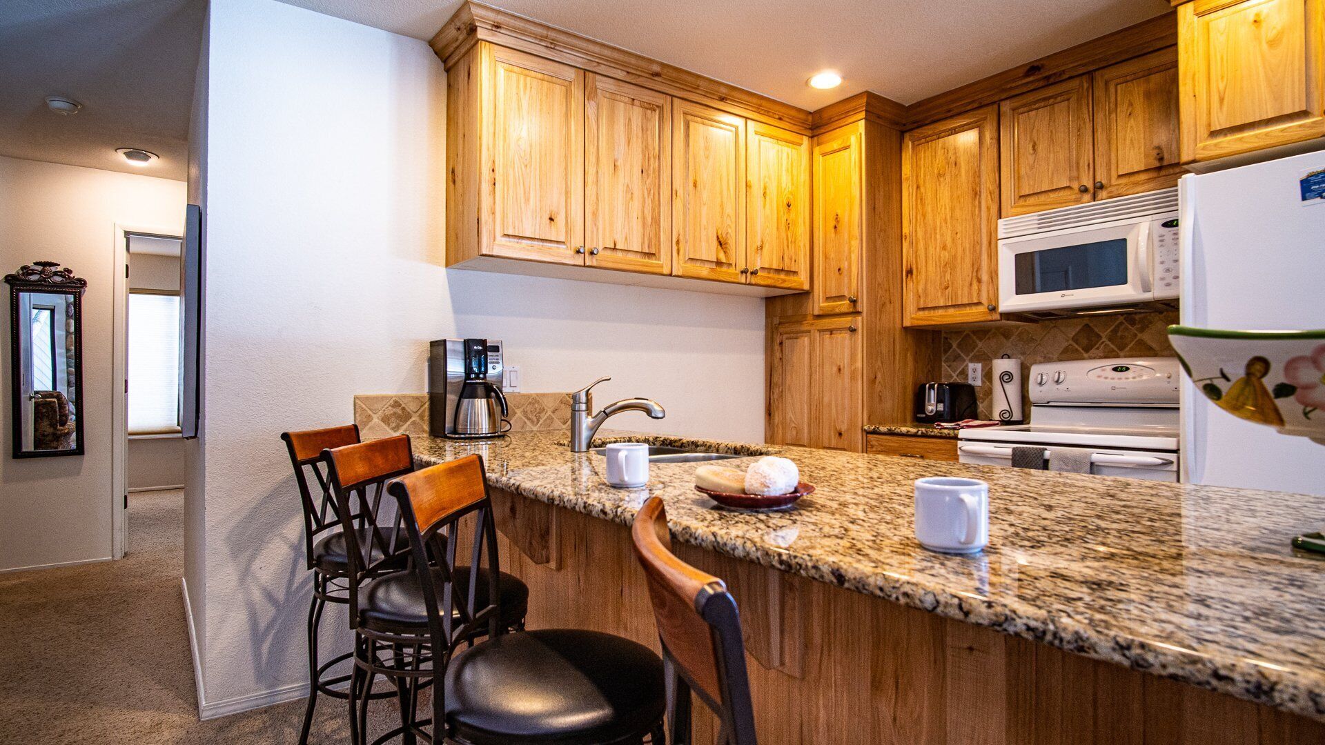 A kitchen with wooden cabinets, a granite countertop, a coffee maker, a microwave, and a refrigerator. There are two bar stools and a sink.