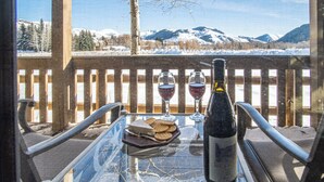 Une table en verre avec une bouteille et deux verres de vin rouge, une assiette de crackers et une vue sur des montagnes enneigées.