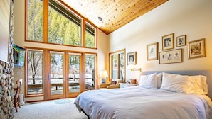 A bedroom with a large bed, wooden ceiling, and multiple windows offering a view of trees. - River House - 4BD Riverfront, Hot Tub, Near SV Ski Lifts! (Ketchum)