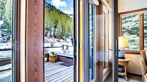 A room with a large sliding glass door leading to a deck with a hot tub, wicker furniture, and a view of a snowy forest. - River House - 4BD Riverfront, Hot Tub, Near SV Ski Lifts! (Ketchum)