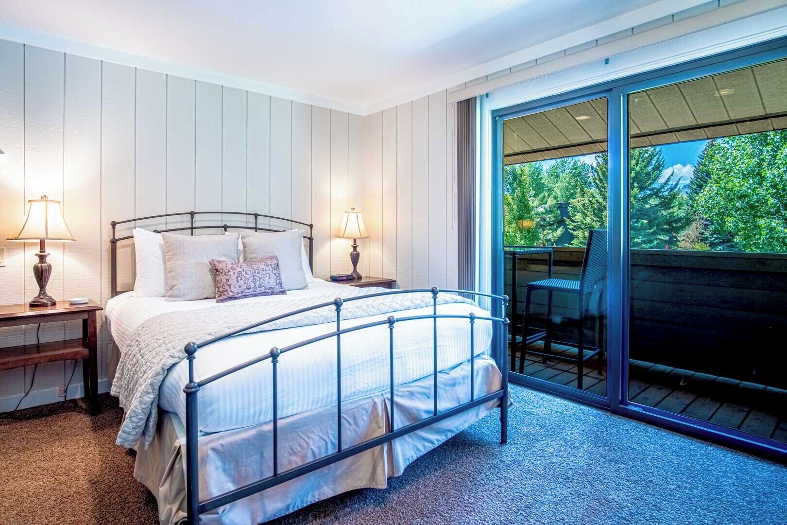 A bedroom with a metal bed frame, two bedside lamps, a large window with a view of greenery, and a sliding glass door leading to an outdoor area.