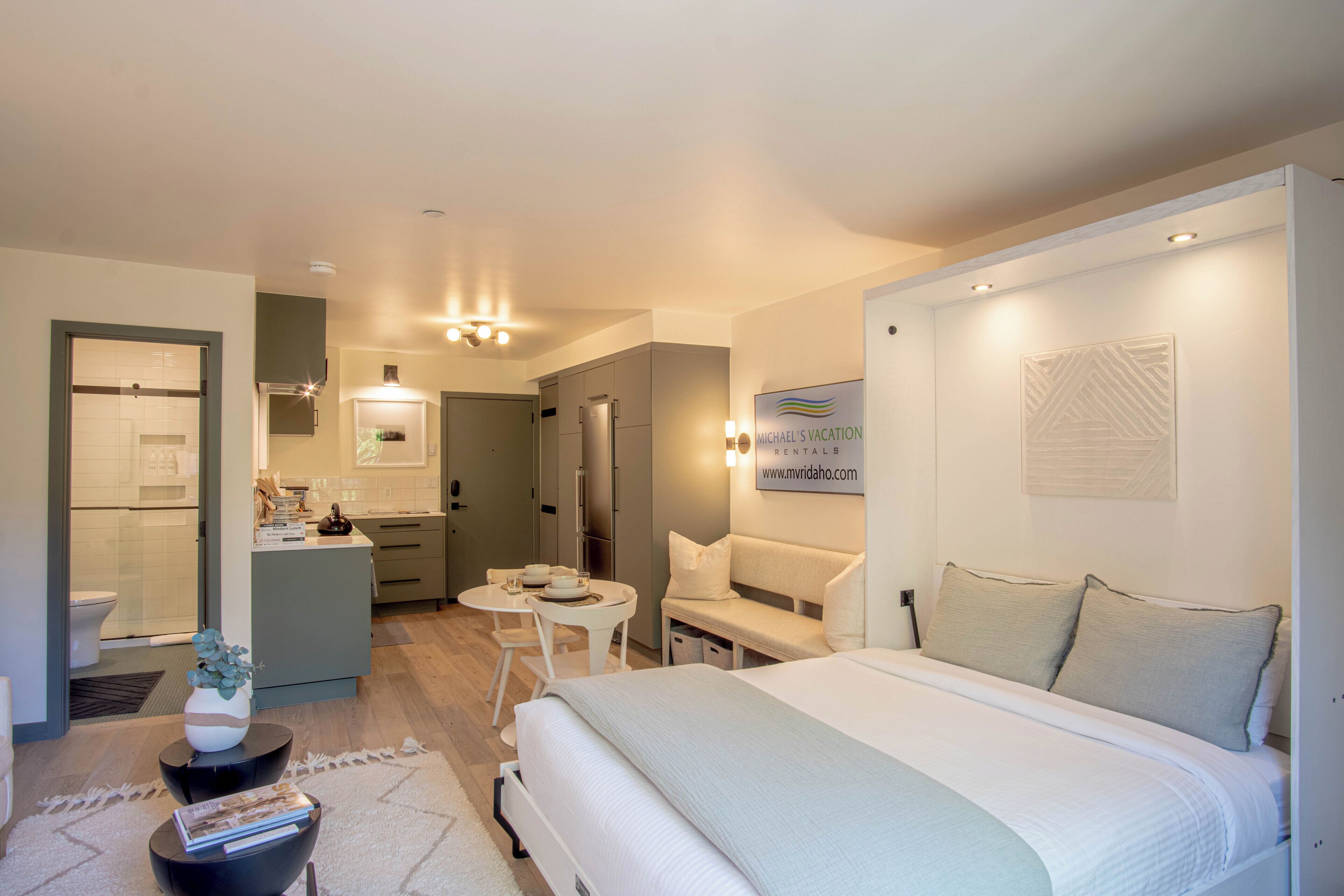 Una habitación de hotel moderna con una cama grande, una zona de estar con sofá y sillones, una pequeña mesa de comedor y una kitchenette.