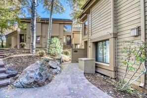 A two-story building with a stone pathway leading to the entrance, surrounded by large rocks and trees. - Snow Creek 1557 - Pool & Hot Tub + Ski-in/ski-out (Sun Valley)