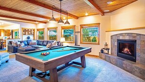 A cozy living room with a pool table, comfortable seating, a fireplace, and large windows offering a view of the outdoors. - Cranbrook Cottage - Family Home + Hot Tub + Pool Table (Hailey)